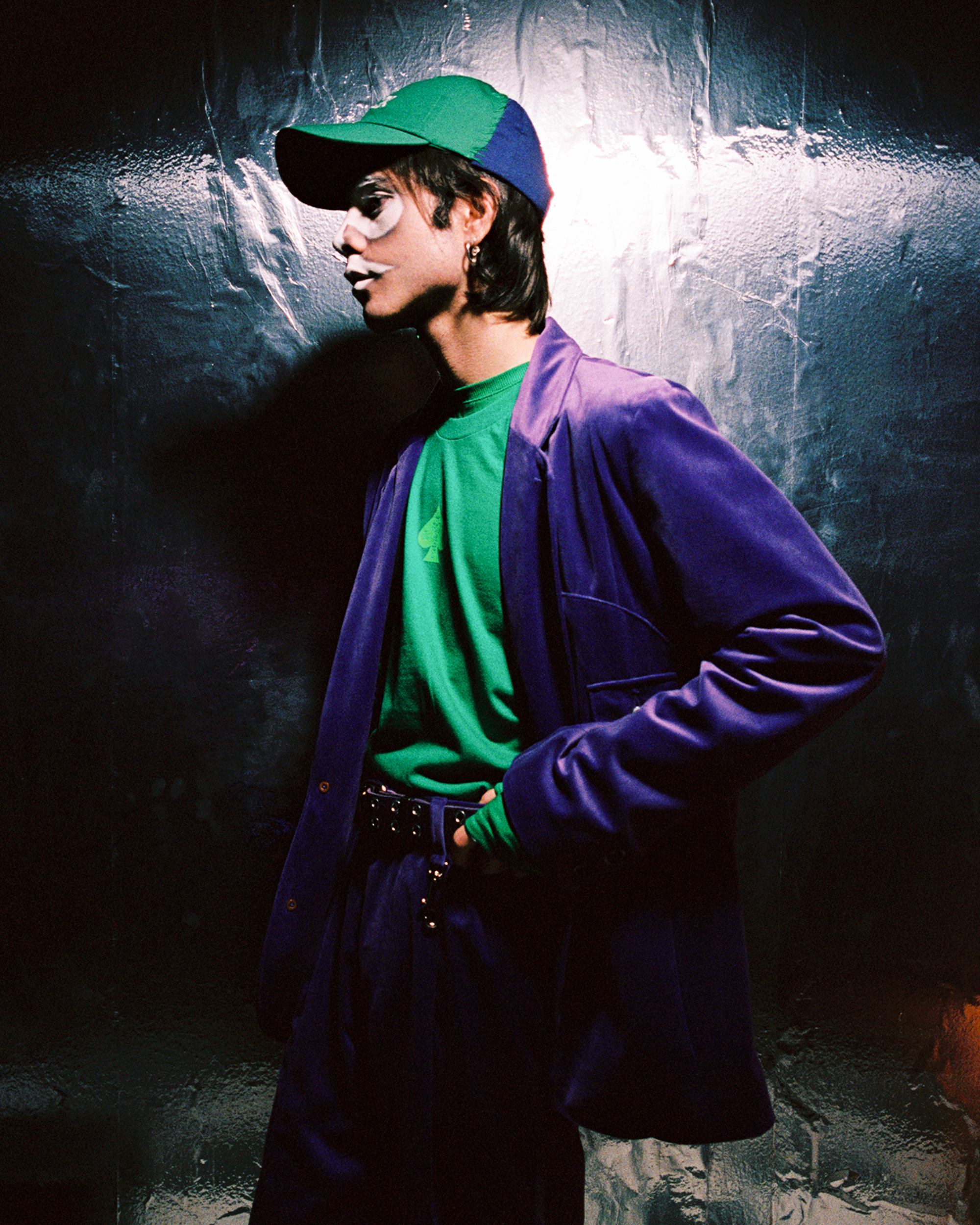 Model standing in profile view with white spade shaped facepaint on against a dark background